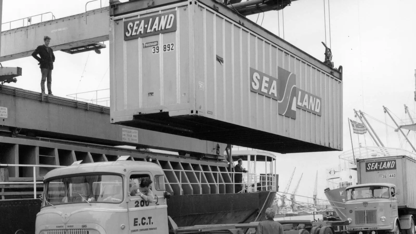 black and white image of a shipping container being unloaded from a ship and onto a truck by a crane in the 1950s